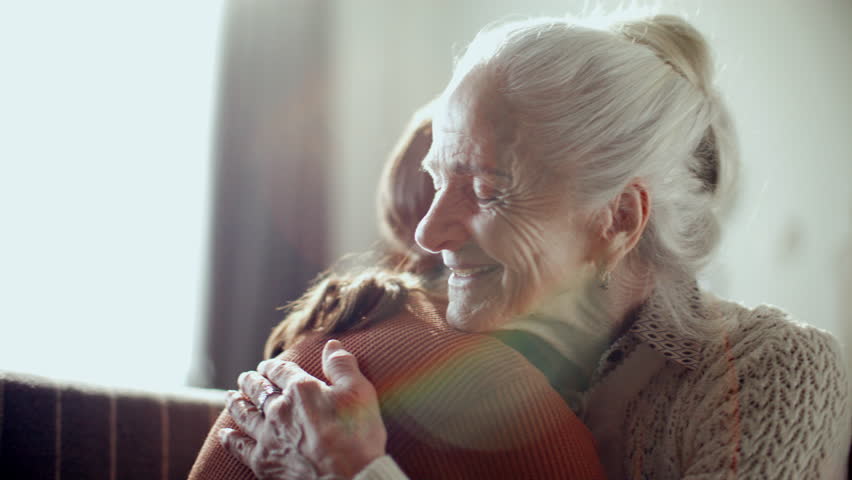 Senior woman hugging young granddaughter, smiling and talking to her on couch in living room filled with sunlight
