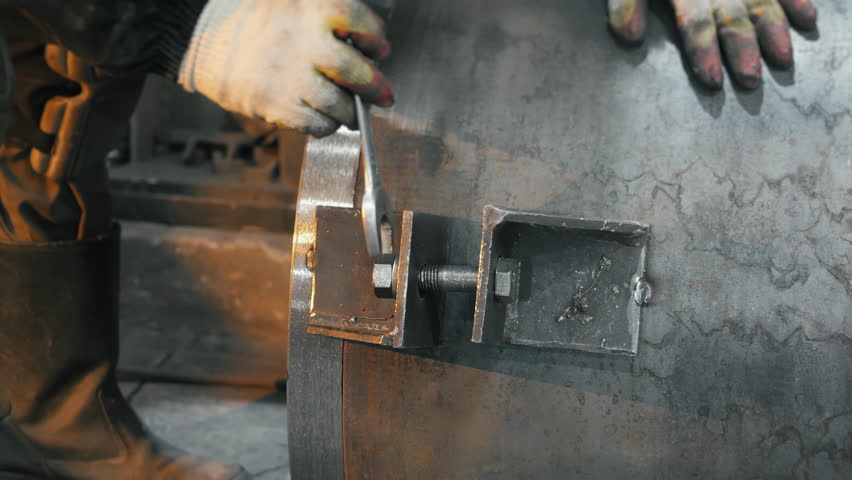 Worker tightens a metal fastener with a wrench on a large steel structure in an industrial workshop. Close up action showing mechanical adjustment, tools, and heavy metal components.