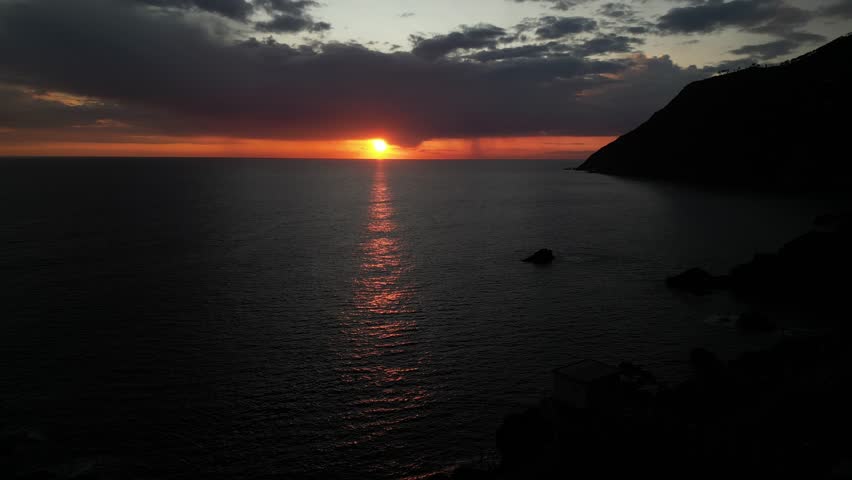 beautiful scenic epic drone flight over the sea at sunset with the red sun setting into the sea on the horizon with a colorful sky full of gray clouds in Framura Italy Liguria landscape 