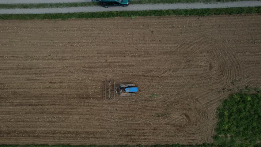 Top view of a blue tractor plowing a dry field next to a green pickup truck parked on a dirt road that separates the plowed field from a green field