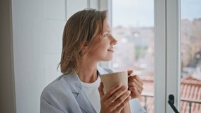 Portrait girl holding mug by window in bright apartment feeling content alone. Smiling lady enjoying tranquil morning posing camera at city view. Happy relaxed woman dream with warm beverage at home - Powered by Shutterstock - Get 15% off with code: PIKWIZARD15