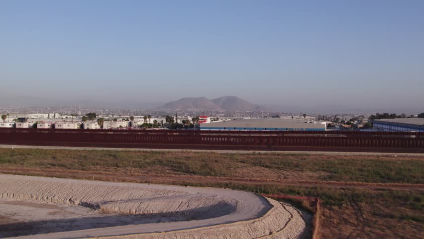 A drone shot captures a wide expanse of terrain, transitioning from freshly cut earthwork to a stretch of red border fencing.