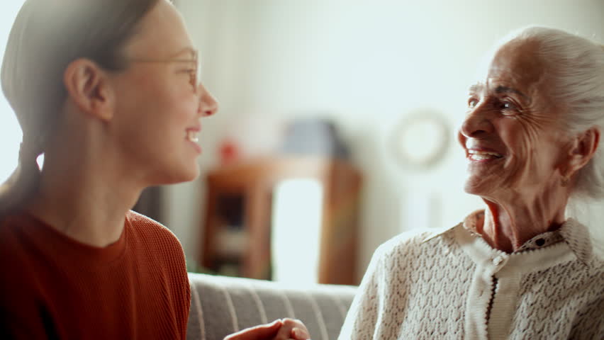 Elderly woman smiling and talking to young granddaughter, enjoying warm conversation on couch in living room filled with bright sunlight