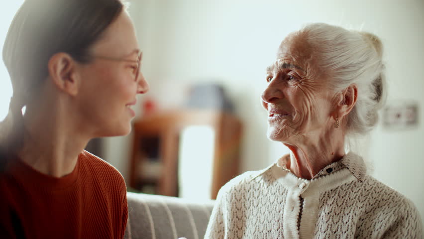 Elderly woman smiling and talking to young granddaughter, enjoying warm conversation on couch in living room filled with bright sunlight