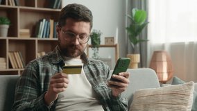 Smiling satisfied Caucasian adult addict man male guy shopping mobile phone at home couch payment online buying internet purchase holding credit card web money cashless e-commerce buy smartphone app - Powered by Shutterstock - Get 15% off with code: PIKWIZARD15