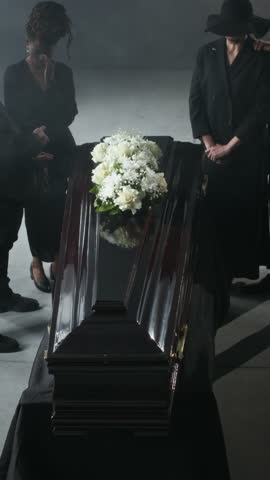 Vertical high angle shot of polished wooden coffin, adorned with white flowers amidst somber gathering of mourners dressed in black at funeral