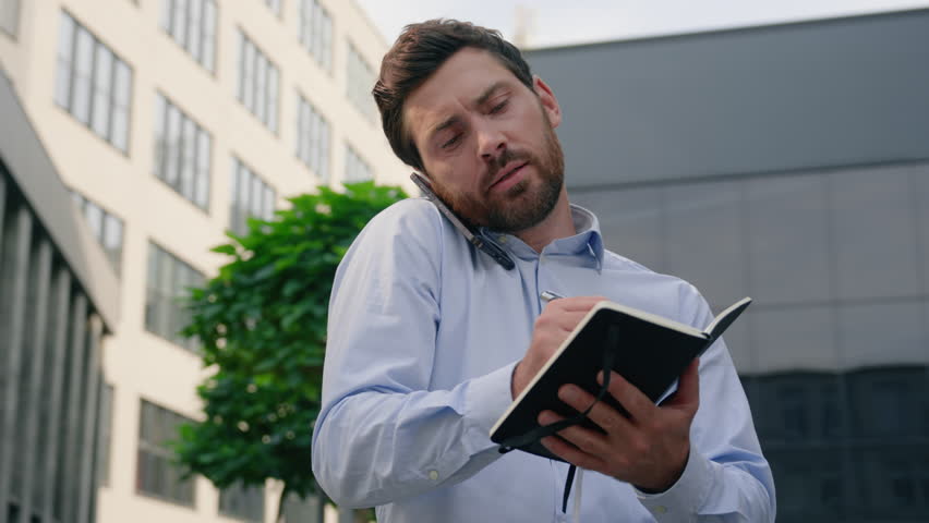 Serious male employer talking mobile phone call in city writing notes outdoors downtown write in notebook outside businessman business talk company negotiate man guy entrepreneur speaking smartphone