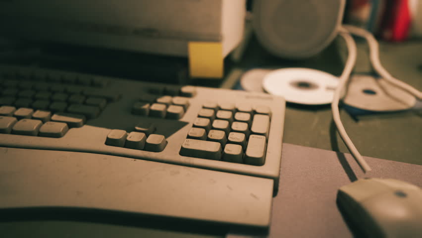 A neglected desk holds a dusty keyboard and mouse alongside scattered CDs and speakers, reflecting a forgotten workspace in a dimly lit room.