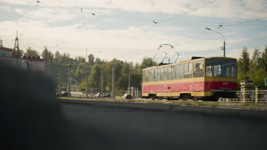 Yellow and red tram moves along city track surrounded by green trees, buildings, and overhead electric wires on clear sunny day, capturing urban transport scene with peaceful suburban atmosphere