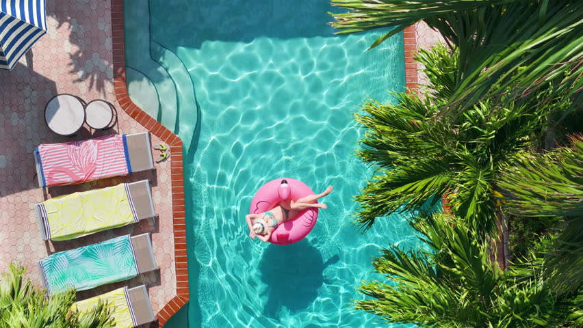 Top-down view of happy carefree woman enjoying vacation stay in luxury tropical villa with pool in Miami, Florida. Sexy female on pink flamingo float on summer trip sunbathing in pool. Luxury travel