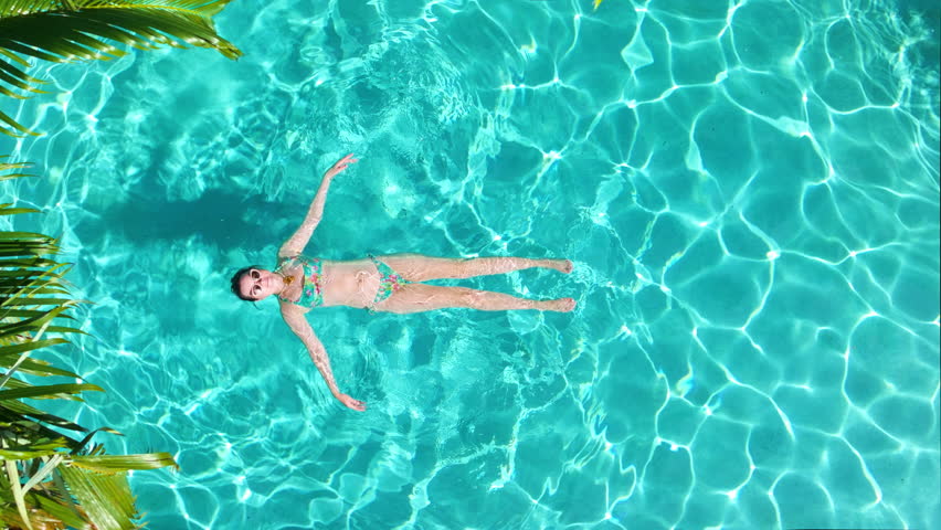 Top-down view of serene tropical swimming pool with a woman floating calmly on her back,arms outstretched in clear turquoise water, captured in slow motion on sunny summer day surrounded by palm trees