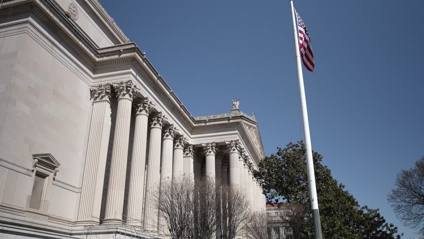 The National Archives building stands prominently with classic architecture, featuring tall columns and an American flag amidst a clear blue sky in Washington DC.