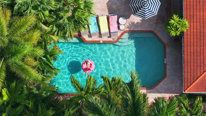 Aerial view of a tropical backyard pool with happy woman relaxing on a flamingo float, surrounded by palm trees, colorful loungers with towels, striped umbrella on sunny summer day. Travel background