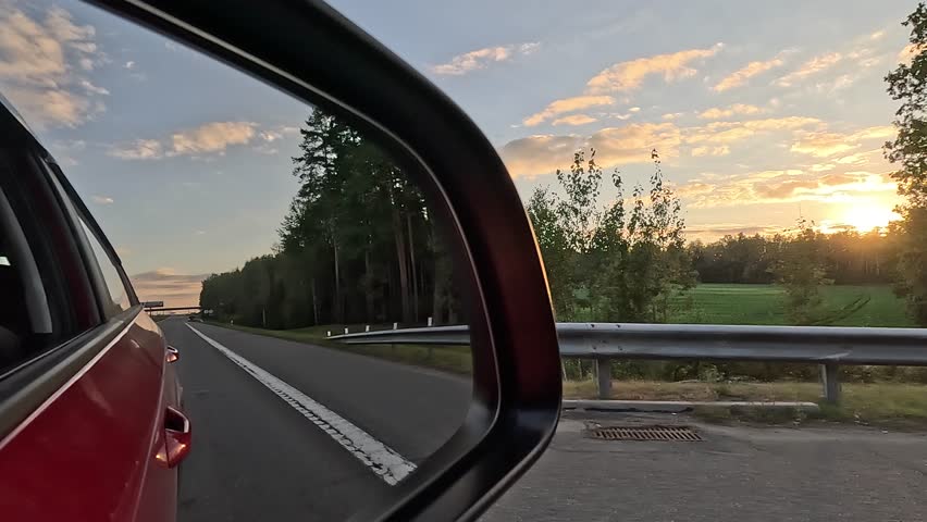 The sun is reflected in the mirror and window, evening road. View in the side rear view mirror of a car driving along the highway, in a red car