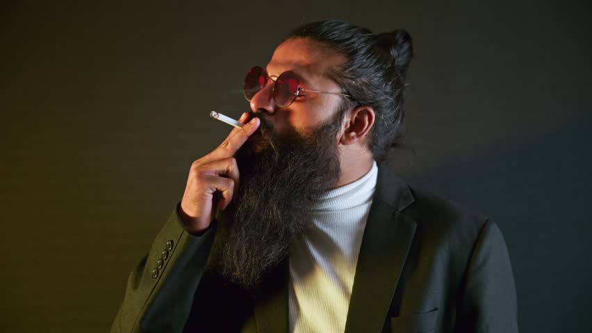 Refined scholar draws thoughtfully on his cigarillo, Man with a beard holds cigarette while standing, Stylish gentleman smoking glowing tobacco roll, Guy in suit enjoy filter stick, Businessman exhale