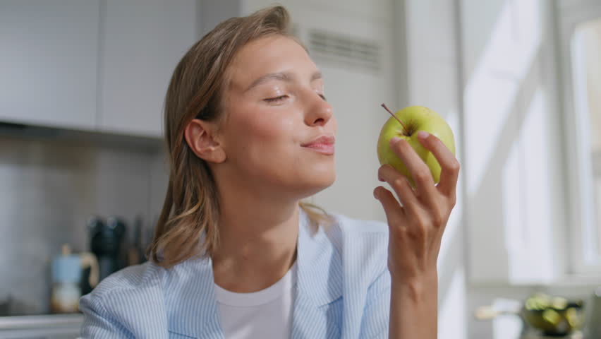 Closeup woman eating apple in modern apartment. Smiling girl tasting biting green fruit enjoying sweet snack in morning. Joyful female savoring fresh organic food. Healthy food nourishment concept.