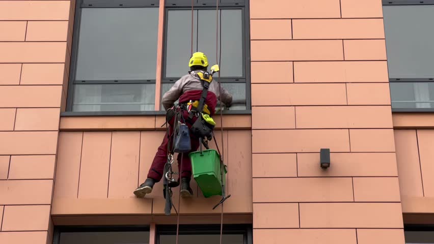 Window cleaner working on a glass facade modern skyscraper. An industrial mountaineering worker washes the glass windows of a high-rise building. A man window cleaner washes the facade of a building. 