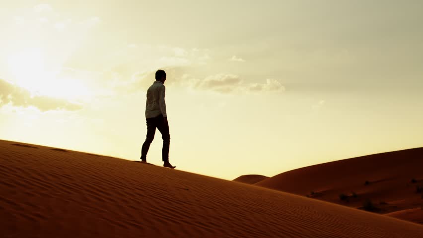 Slow motion video: Silhouette of a human walking on the sands in the desert in windy weather while sunrise.  Young man in stylish clothes walks barefoot on the sand dunes in the desert at sunset. 