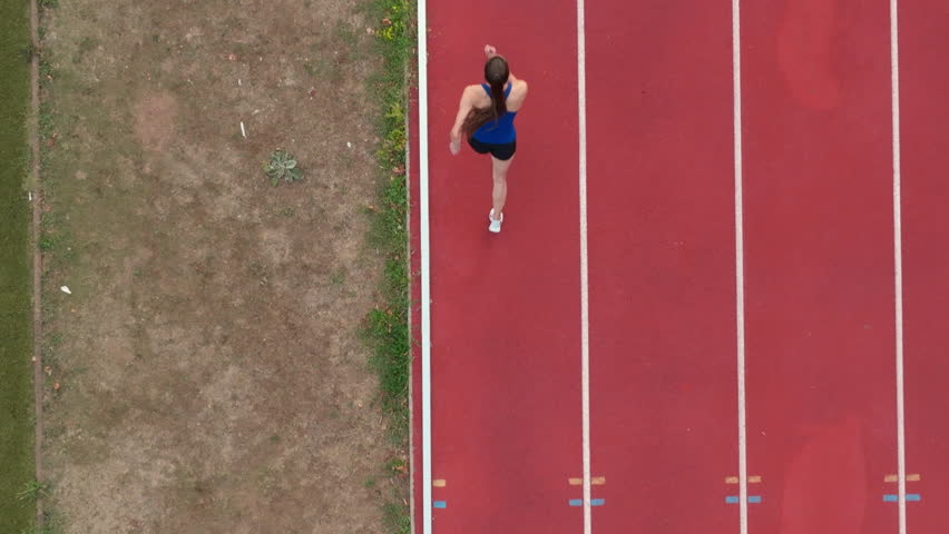 Top down view aerial drone shot of a professional female athlete sprinting on a large stadium athletics track. Dynamic slow motion cinematic footage captures her power, speed, and determination in