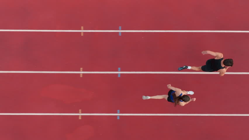 Aerial drone shot from above of an athletic couple jogging side by side on a stadium track, captured in slow motion, emphasizing their fitness, rhythm, and energy as they move in perfect sync