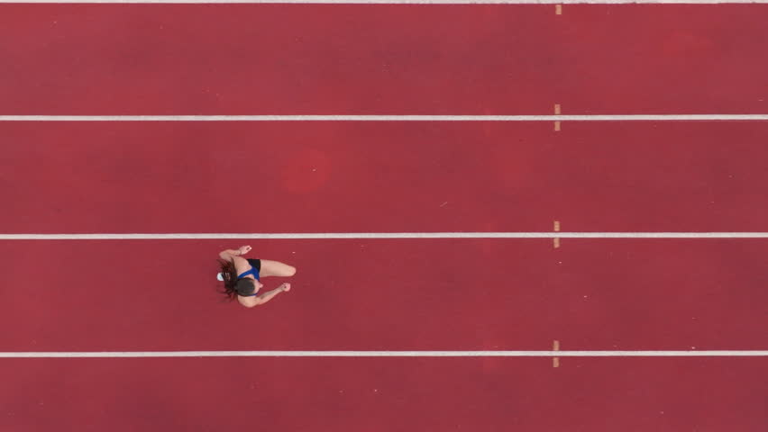 Top down view aerial drone shot of a professional female athlete sprinting on a large stadium athletics track. Dynamic slow motion cinematic footage captures her power, speed, and determination in