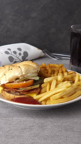 Close-up of a plate with a burger, straight and curly fries. A hand picks up a fry and dips it in tomato ketchup. Delicious fast food moment. Vertical stock video for food and dining visuals.