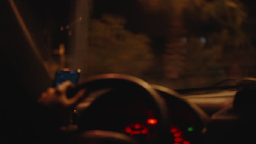 A Driver's Hand Holding the Steering Wheel with a Smartphone Navigation Screen Visible, Illuminated Dashboard Lights, and a Blurred Nighttime View Through the Windshield. Night Driving Focus