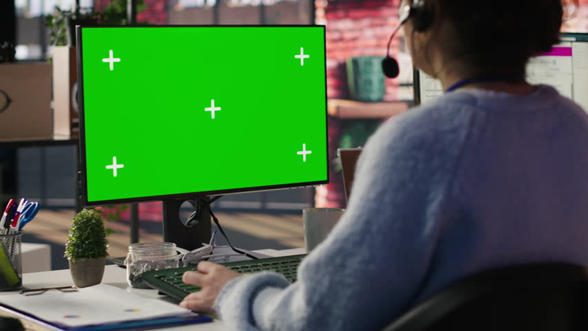 Call center employee working on green screen mockup PC, doing account management in office. Customer support agent in front of chroma key isolated screen computer handling calls, camera A close up