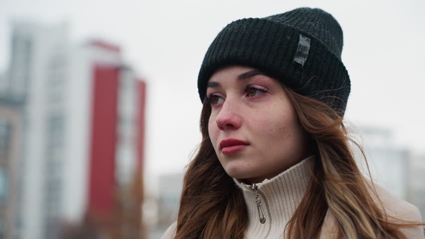 Close shot of thoughtful girl in black cap and brown jacket walking with serious expression and wind-blown hair against blurred city background under overcast sky during quiet cold autumn day