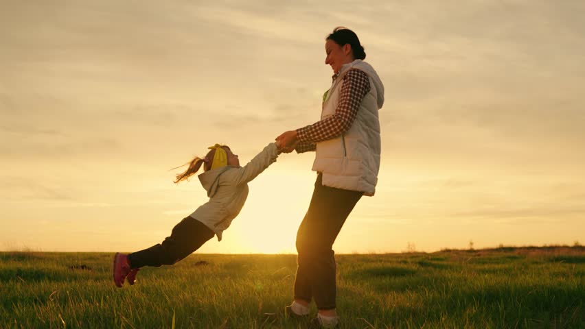 Child having fun in flight. Family weekend nature. Mother spins little daughter on green grass, sunset. Happy family playing on lawn. Baby daughter play with mom, kid fly, mother holding child hands