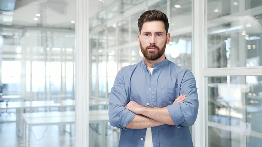 Portrait of serious successful businessman standing with crossed arms at workplace in a business office. Confident handsome bearded manager looking at camera. Proud entrepreneur posing in workspace