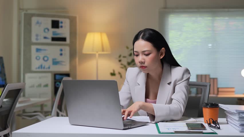 Fatigued asian businesswoman working late at night, drinking coffee while struggling to stay focused and complete laptop tasks in dimly lit corporate office setting