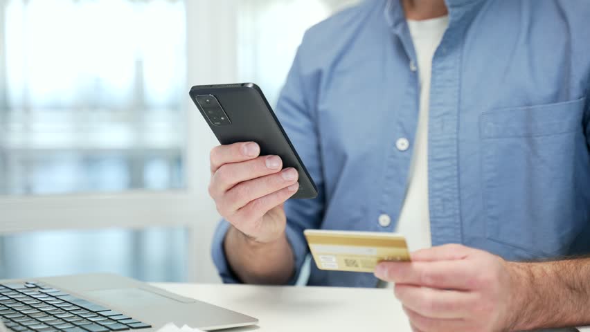 Close up of male hands using phone to make online transaction with credit card at desk in business office. Shopper makes digital payment, pays bills, spends money on internet, e-commerce, e-shopping