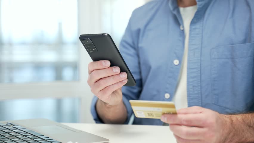 Close up of male hands using phone to make online transaction with credit card at desk in business office. Shopper makes digital payment, pays bills, spends money on internet, e-commerce, e-shopping