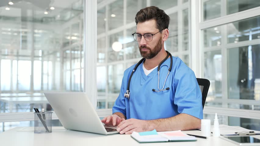 Confident doctor in blue coat typing on laptop sitting at workplace in modern hospital office. Focused handsome medical worker physician is engaged in work or chats online with patient using computer