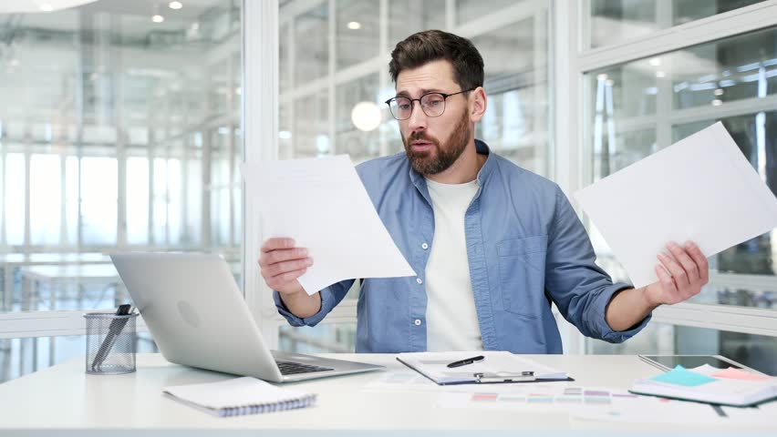 Confused disappointed businessman having difficulty with paperwork sitting at desk at workplace in modern business office. Puzzled entrepreneur looks at documents and cannot understand the problem