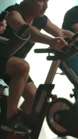 Sportmen training the muscles of the legs on the exercise bicycles in the gym in a row. Healthy lifestyle concept, the desire to preserve youth. Final focusing on pretty lady. Vertical shot.