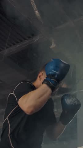 Young professional athlete strikes a punching bag aggressively with great force while wearing gloves. Smoky gym with lots of exercise equipment. Vertical shot.