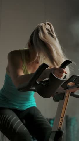 Pretty sexy girl in leggings and a T-shirt pedalling on a bike training apparatus. Cardio hard training for a handsome woman in a smoky gym. Slow motion handheld zooming shot. Vertical shot.