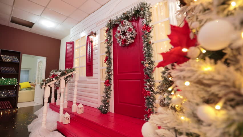 Approaching red door with a festive wreath and garland around. Low angle view at the porch decorated for Christmas.
