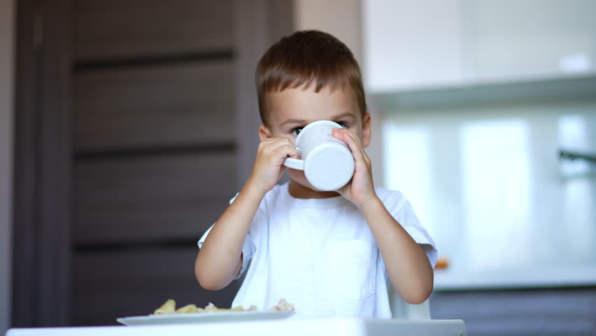 Toddler boy in white t-shirt drinks from a cup. Kid finishes his drink and gives a cup to his mom.