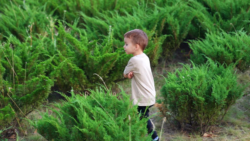 Little boy walks with hands folded on the chest. Frowning kid in the park among the greenery.