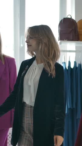 Young female friends coming closer to clothes rack and starting to choose new apparel while visiting shop together. Vertical shot.