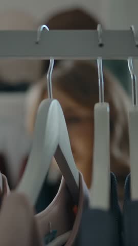 Blurred female customer walking behind clothes rack and choosing new garment while visiting store in weekend. Vertical shot.