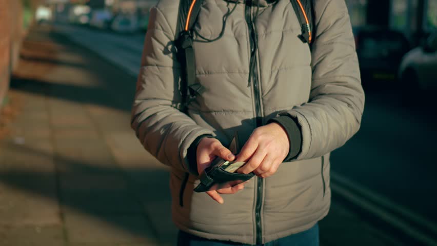 Man counting money dollars in wallet while standing on street. Male checking cash outdoors in quiet neighborhood. Guy holding wallet and organizing bills near roadside area