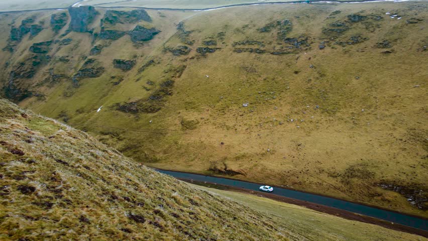 Car traveling on mountain road with steep grassy slopes. Vehicle driving through scenic route in Peak District, United Kingdom. White car moving along winding valley road surrounded by rugged terrain