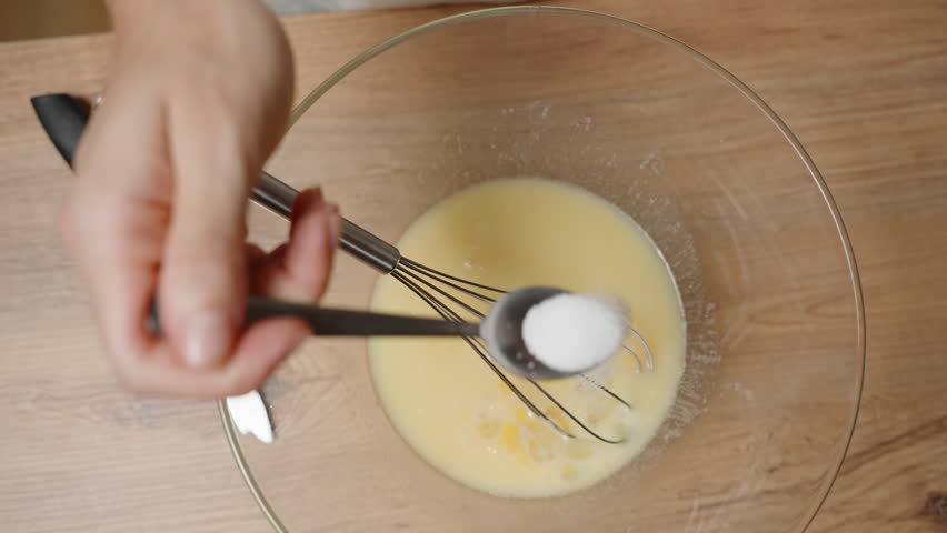 Person Adding a Spoonful of Salt into Egg Mixture in a Glass Bowl, Preparing Ingredients, Focus on Baking Process. Slow motion.