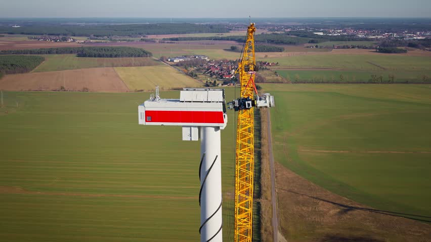 Droneshot of wind turbine nacelle during gearbox installation by crane in a rural landscape on a clear day, movement drone right.