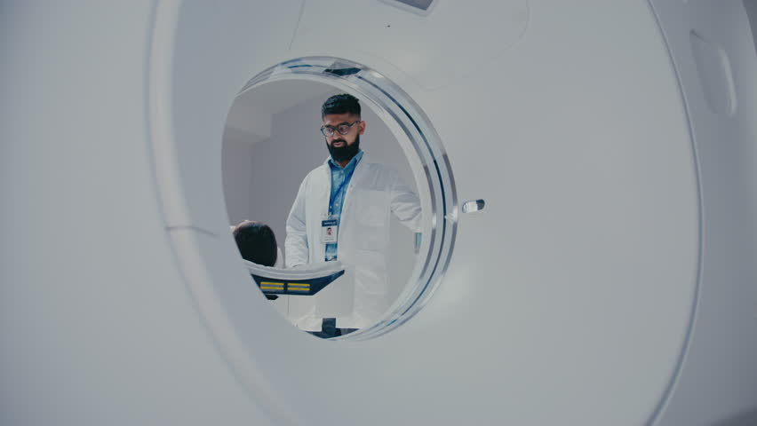 Serious healthcare technician monitoring scanning phase while woman positioned on MRI bed with red laser lines projecting. Conducting advanced imaging process inside bright hospital facility.