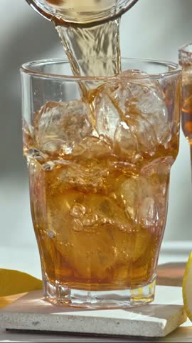 fresh  iced tea flowing in glass with ice  from glass carafe on sunny white wooden table top in slow motion
